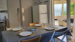 A dining area with a table set for four, including plates, glasses, and cutlery. There are two chairs, a refrigerator, and a window with a view of greenery.