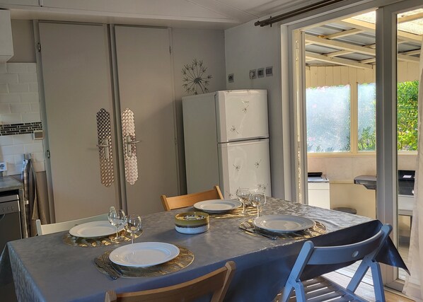 A dining area with a table set for four, including plates, glasses, and cutlery. There are two chairs, a refrigerator, and a window with a view of greenery.