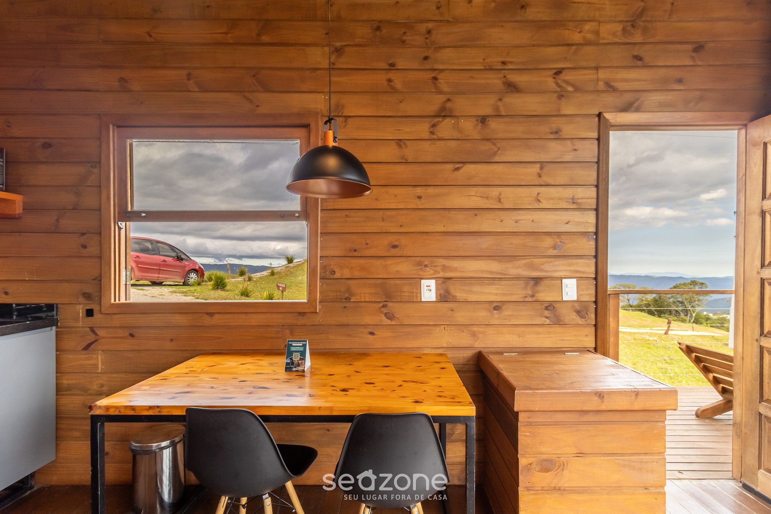 A wooden cabin interior with a dining table, chairs, a hanging lamp, and a window overlooking a red car and landscape.