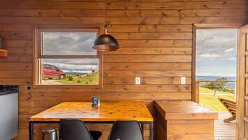 Interno di una baita in legno con tavolo da pranzo, sedie, lampada a sospensione e finestra vista su un'auto rossa e il paesaggio.