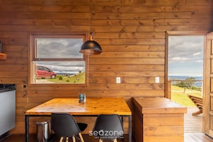 Interior de una cabaña de madera con una mesa de comedor, sillas, una lámpara colgante y una ventana que da a un coche rojo y un paisaje.