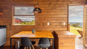 Interieur van een houten hutje met een eettafel, stoelen, een hanglamp en een raam met uitzicht op een rode auto en het landschap.