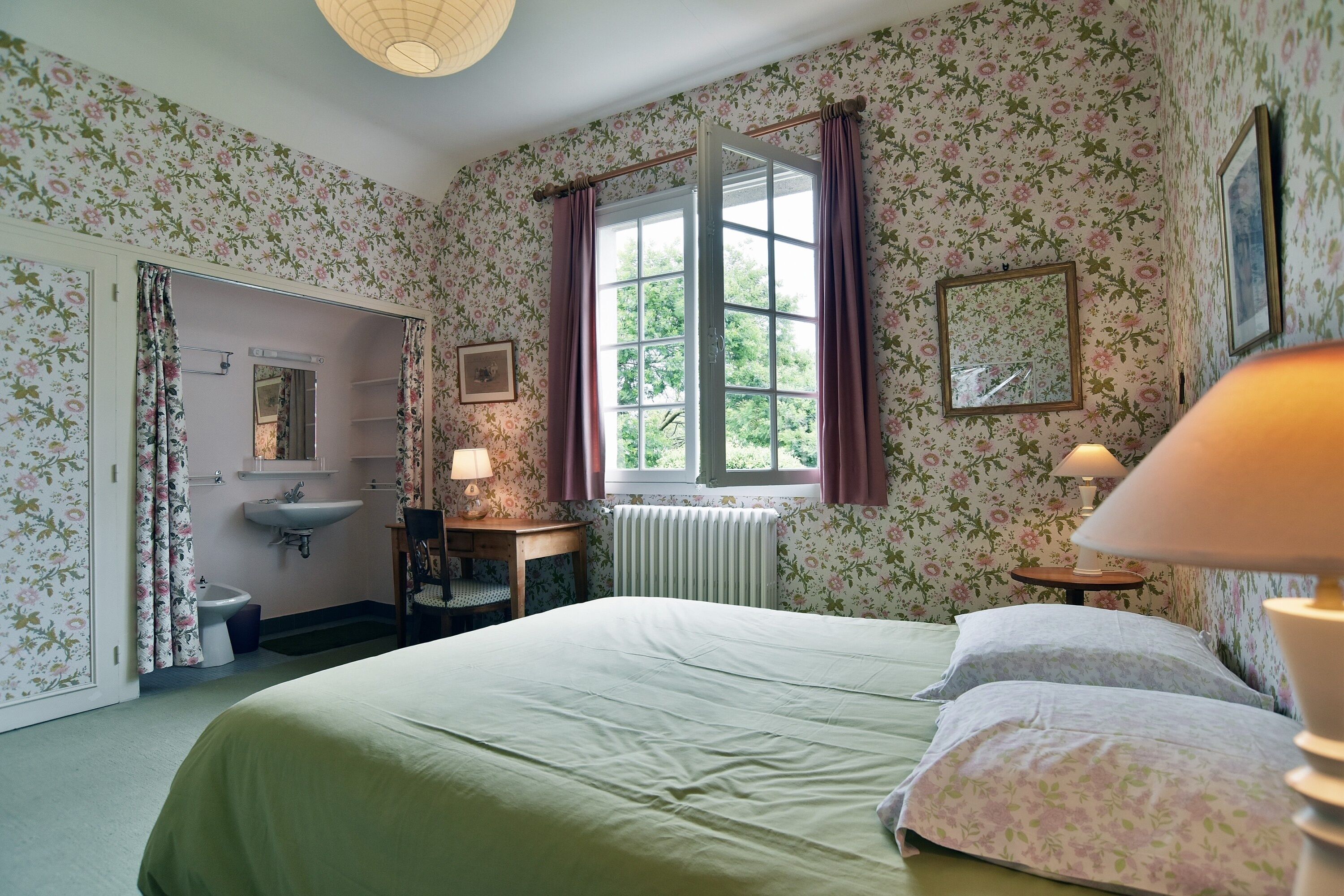 Un dormitorio con papel tapiz floral, una cama con ropa de cama verde, una ventana con cortinas moradas, un radiador y una mesita de noche con una lámpara.