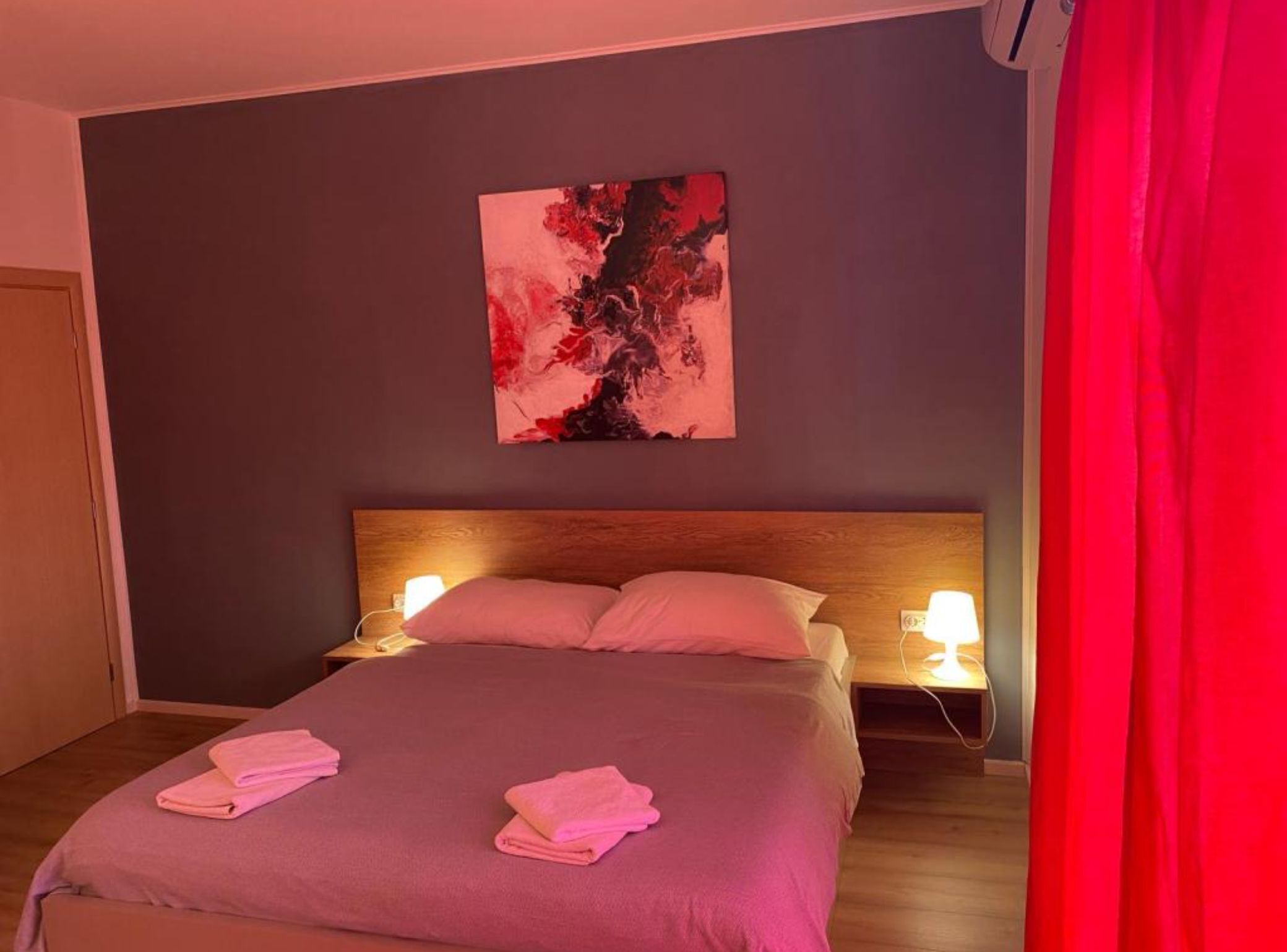 Ein Hotelzimmer mit einem Bett, zwei Nachttischlampen, einem Wandbild im abstrakten Stil und einem roten Vorhang.