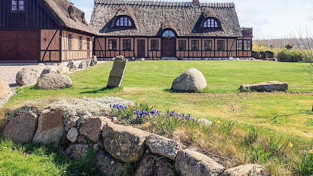 Un bâtiment traditionnel en pierre, doté d’un toit de chaume, d’un mur en pierre et d’un champ herbeux parsemé de fleurs.