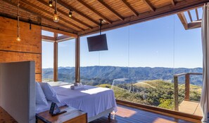 Une chambre à coucher comprenant un lit, une table de chevet, un téléviseur et une grande fenêtre donnant sur la montagne.