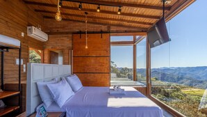A bedroom with a bed, pillows, a bedside table with a remote control, and a window with a view of the landscape.