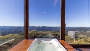 Un bain à remous sur une terrasse en bois, offrant une vue sur un paysage montagneux grâce à de grandes fenêtres.