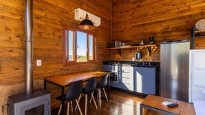 A compact kitchen with wooden walls, a dining area with chairs, a stove, and a refrigerator. - Cabana vista deslumbrante serra VST020 (Anitápolis)
