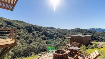 A wooden deck with a brick fire pit and seating, overlooking a lush valley and mountains.