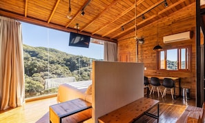 A wooden cabin with a bed, a TV, and a dining area with chairs. - Cabana vista deslumbrante serra VST020 (Anitápolis)