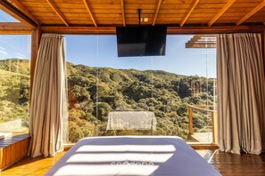 A bedroom with a large bed, wooden ceiling, and a TV. It has a view of a forested hillside. - Cabana vista deslumbrante serra VST020 (Anitápolis)