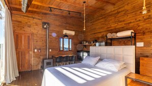 A wooden cabin bedroom with a large bed, a dining table, chairs, and a kitchen area. - Cabana vista deslumbrante serra VST020 (Anitápolis)