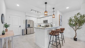 A modern kitchen with white cabinetry, stainless steel appliances, a central island with bar stools, pendant lighting, and a potted plant. - RIVER FLY IN: Garden View (Merritt Island)