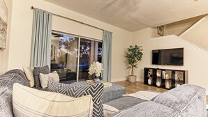 Una sala de estar con un sofá gris, un televisor de pantalla plana, una planta en maceta y un ventanal corredizo que da a un balcón.