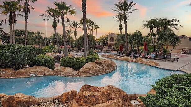 Zona de piscina con palmeras, sillas de descanso y un edificio al fondo.