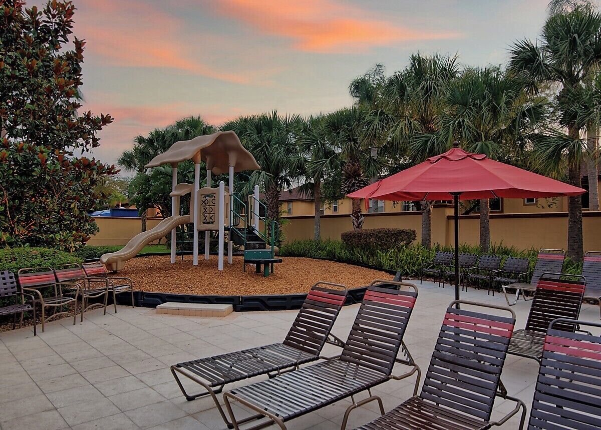 A playground with a slide and climbing structure, surrounded by palm trees and outdoor seating with umbrellas.
