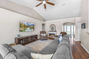 A spacious living room with a large gray sectional sofa, a fireplace, a ceiling fan, and a mounted TV. - Garden view retreat w/ balcony & BBQ grill (Merritt Island)