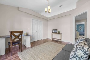 A spacious living room with a wooden floor, a glass coffee table, a dark wooden chair, a patterned sofa, and a small console table with a picture frame.