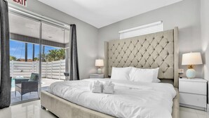 Una moderna camera d'hotel con un letto grande, due comodini con lampade, una porta vetrata che si apre su uno spazio esterno e vista su palme.