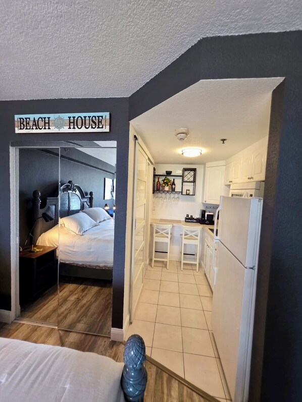 A compact room with a bed, a small desk, and a kitchenette. A sign above the bed reads "BEACH HOUSE".