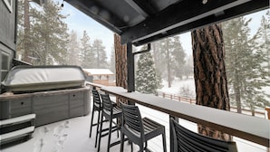 A covered outdoor seating area with a bar, chairs, and a view of snow-covered trees.