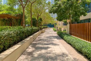 A pathway flanked by neatly trimmed hedges and trees, leading towards a modern building with a wooden fence.