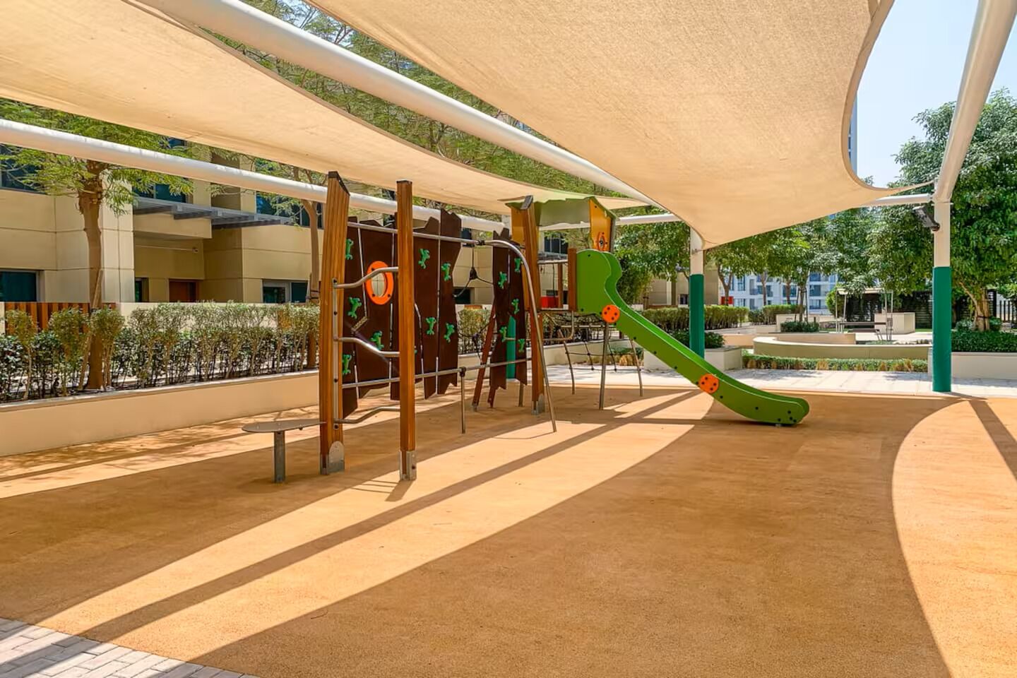 Un patio de juegos al aire libre con estructuras para escalar y un tobogán, rodeado de una zona con sombra y un jardín ajardinado.