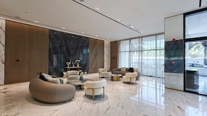 A modern lobby with marble flooring, a seating area with sofas and armchairs, and large windows offering a view of the outside.