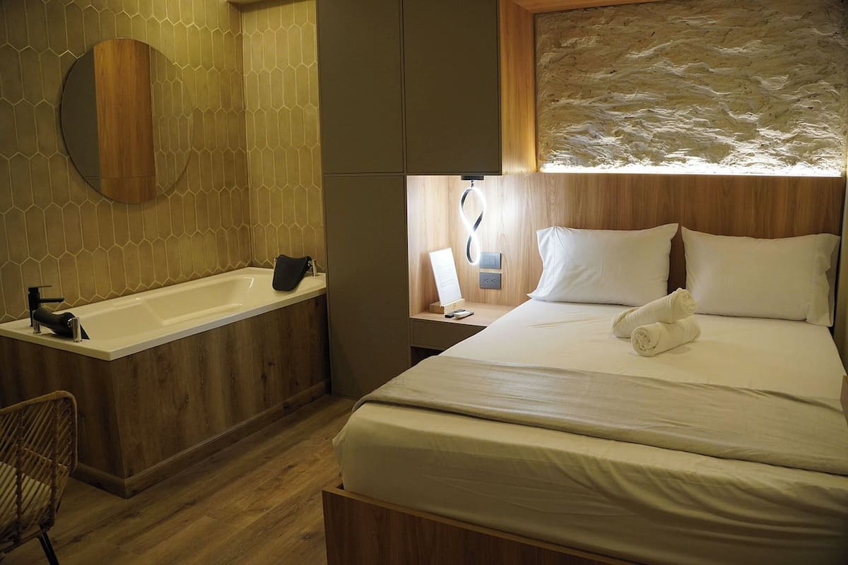 A modern hotel room with a wooden floor, a large bed with white linens, a round mirror, a bathtub, and a nightstand with a lamp.