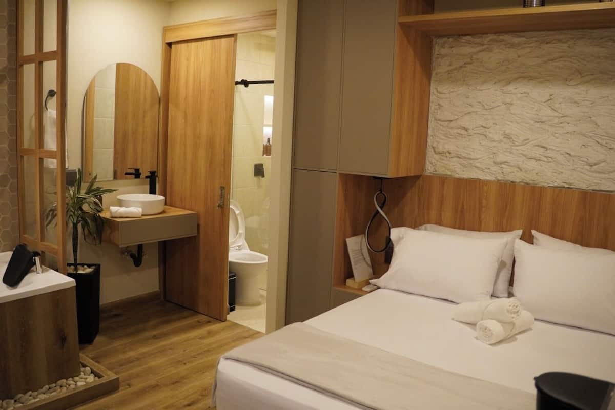 A hotel room with a wooden floor, a bed with white linens, a wooden vanity with a sink, and a mirror.