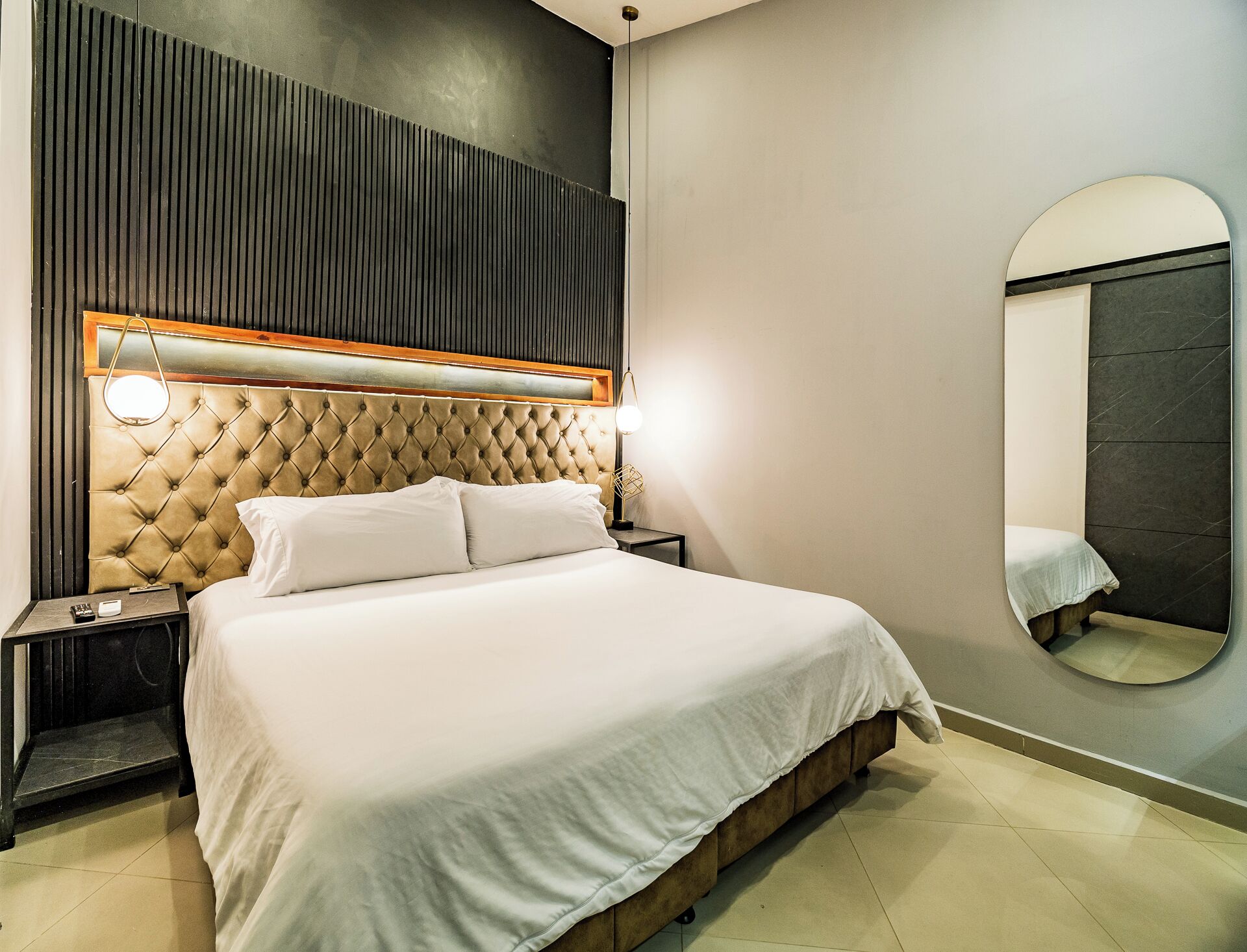 A bedroom with a tufted headboard, a bedside table with a lamp, a large mirror, and a bed with white bedding.