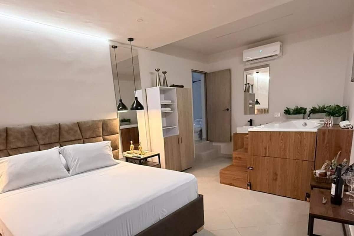 A modern bedroom with a large bed, a wooden dresser, and a small table with a lamp.