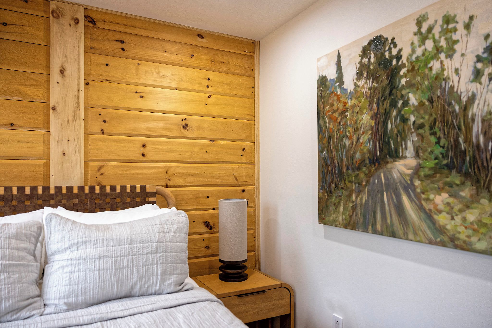 Un dormitorio con cabecera de madera, una mesita de noche con lámpara y un cuadro de un sendero en el bosque.