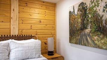 Un dormitorio con cabecera de madera, una mesita de noche con lámpara y un cuadro de un sendero en el bosque.