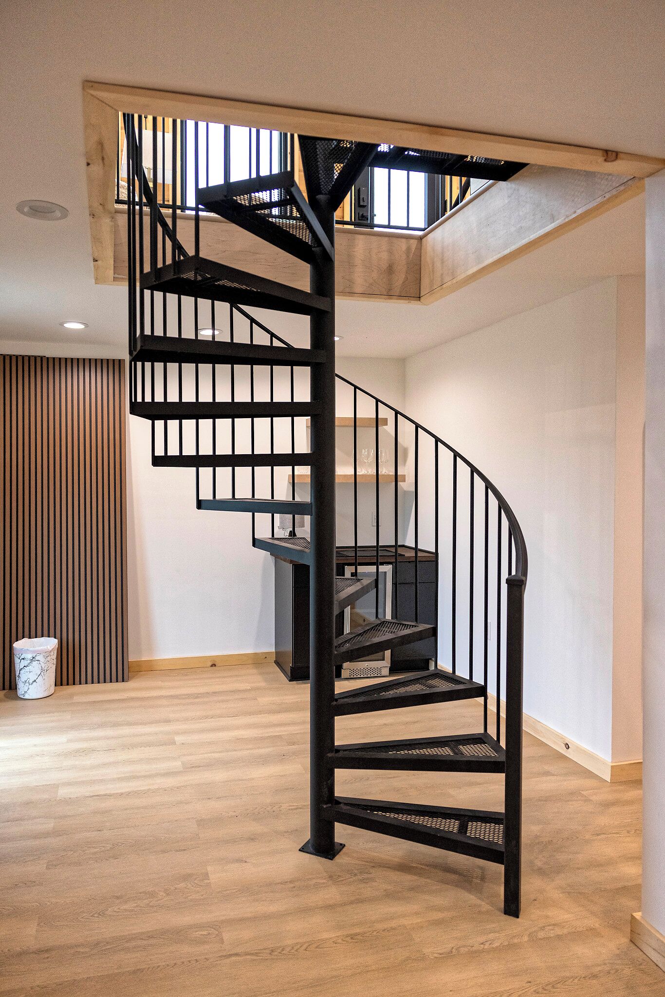 A modern spiral staircase with black railings in a room with wooden flooring and a sloped ceiling.