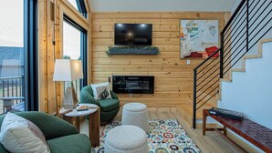 Una sala de estar acogedora con un sofá verde, una mesita, un televisor montado en la pared y una escalera.