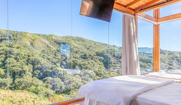 A bedroom with a large window overlooking a forested hillside. - Incrível cabana vista da serra VST021 (Anitapolis)