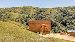 A wooden cabin with a wooden staircase leading to a door, situated on a grassy hill with a forested background. - Incrível cabana vista da serra VST021 (Anitapolis)
