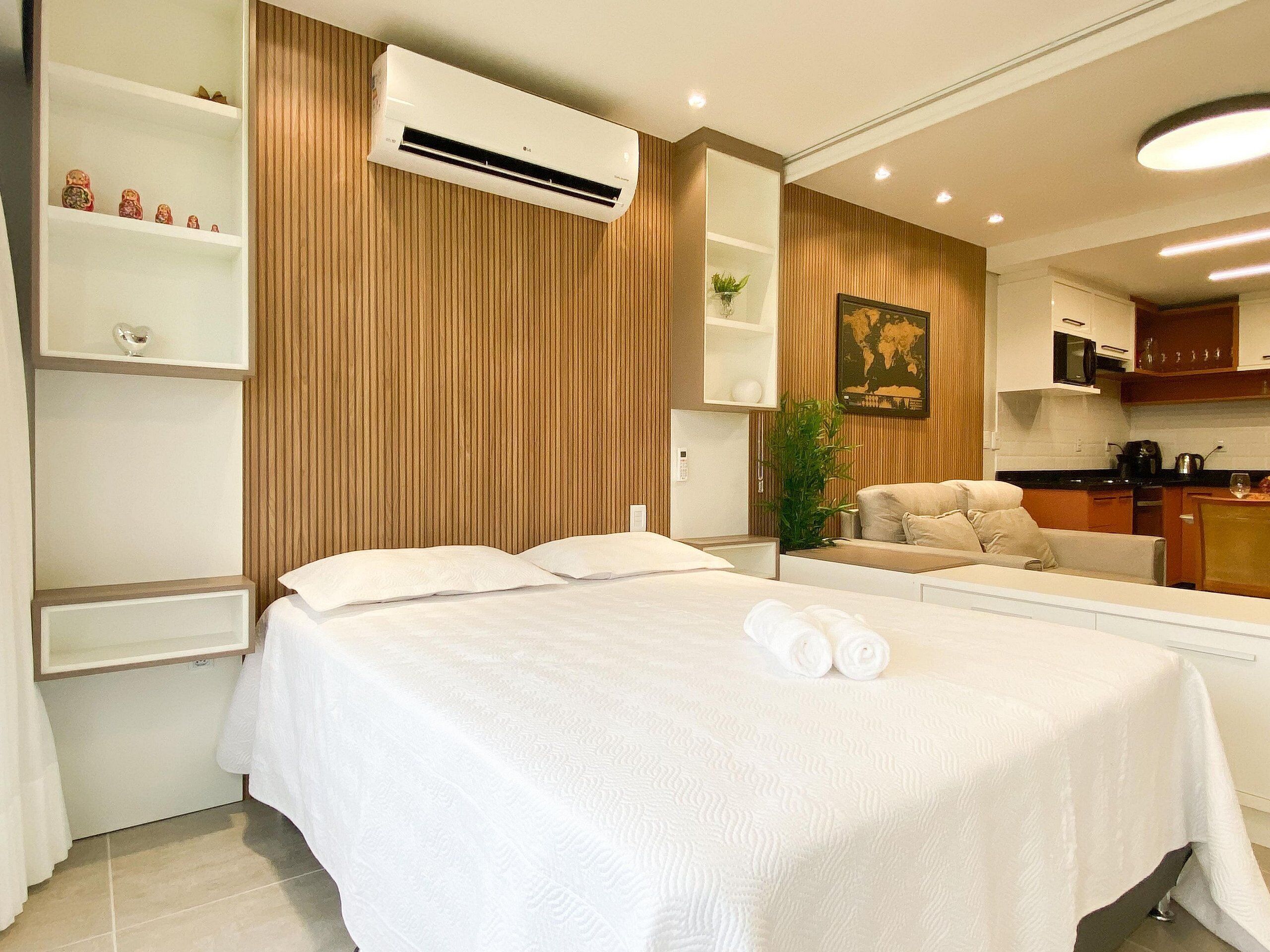 A hotel room with a bed, a small kitchenette, a wooden panel wall, and a mounted air conditioner.