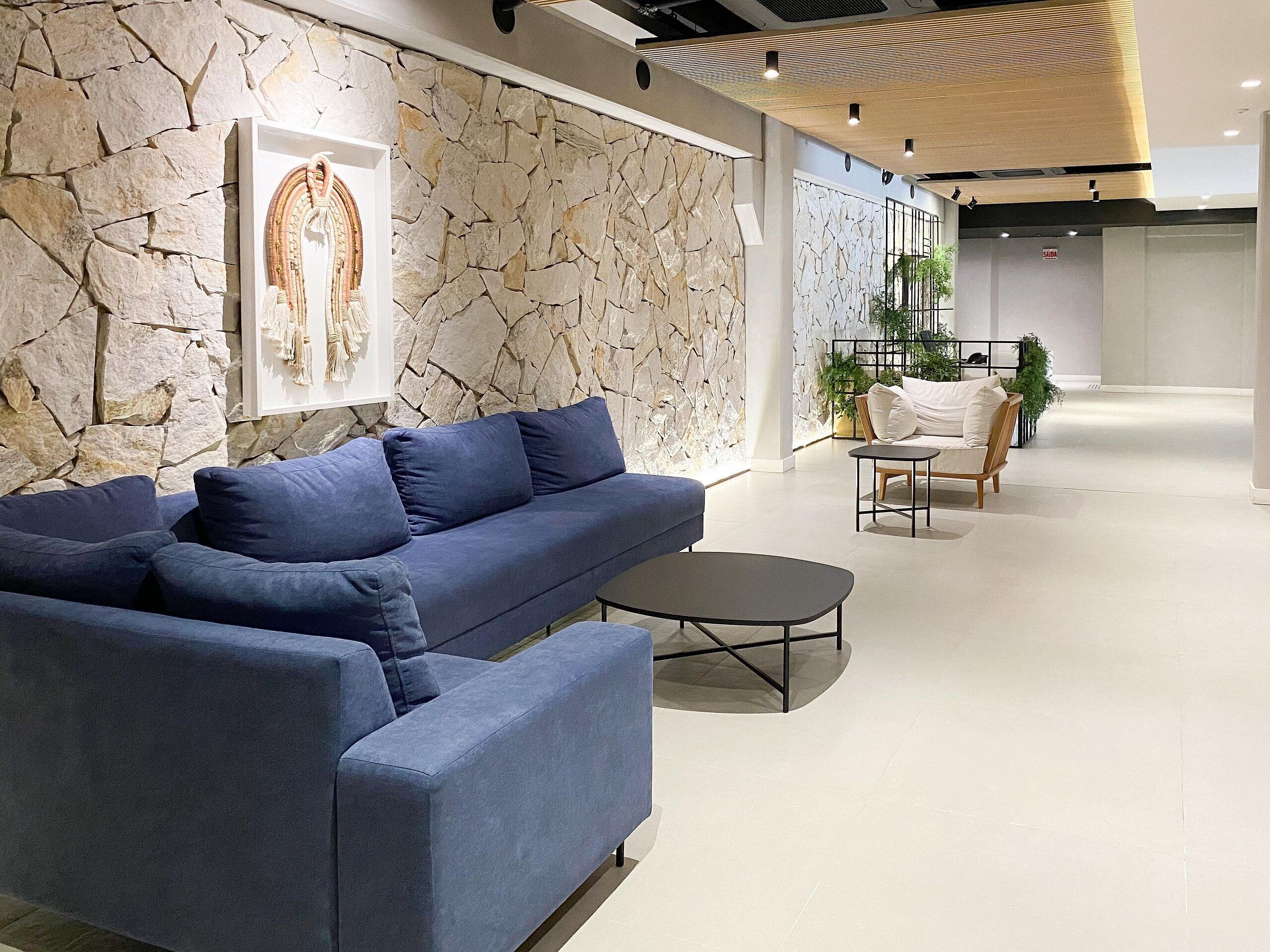 A modern lounge area with a blue sofa, a small round table, and a chair. There is a stone wall with a framed artwork and a plant in the background.