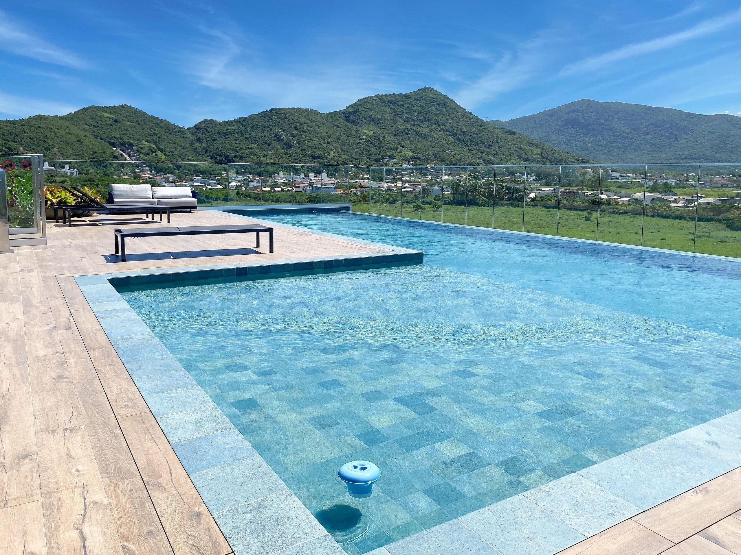 Piscina com água azul cristalina, rodeada por um deck de madeira e uma área de descanso com cadeiras brancas.
