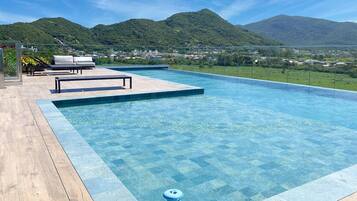 Piscina com água azul cristalina, rodeada por um deck de madeira e uma área de descanso com cadeiras brancas.