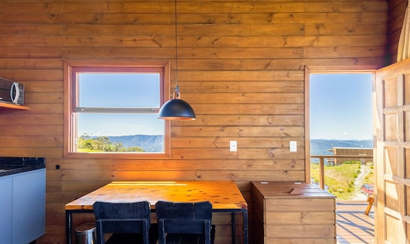A wooden interior with a table and chairs, a window with a view of mountains, and a pendant light.