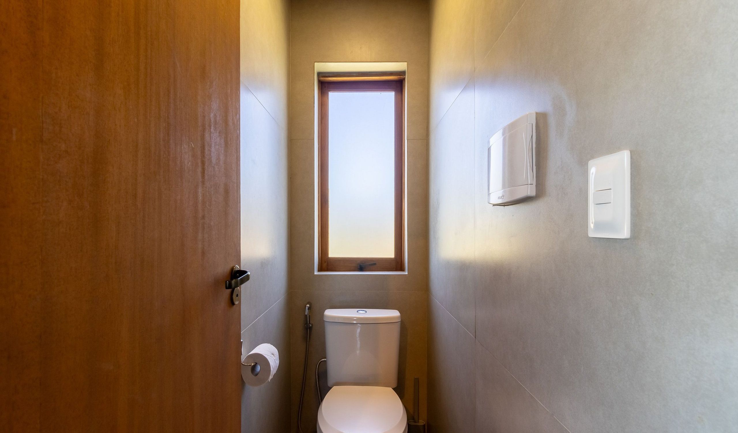 A narrow bathroom with a toilet, a small window, and a wall-mounted heater.