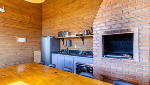 A compact kitchen with wooden walls, a brick oven, a refrigerator, and a dining table with chairs. - Cabana vista panorâmica e jacuzzi VST022 (Anitápolis)