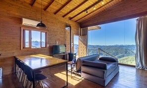 Interior de madera con comedor, sofá y vistas a montañas a través de amplios ventanales.