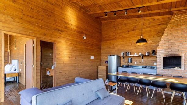 A cozy cabin interior with a wooden interior, a dining area with a long table and chairs, a kitchen with a brick oven, and a seating area with a sofa and cushions. - Cabana vista panorâmica e jacuzzi VST022 (Anitápolis)