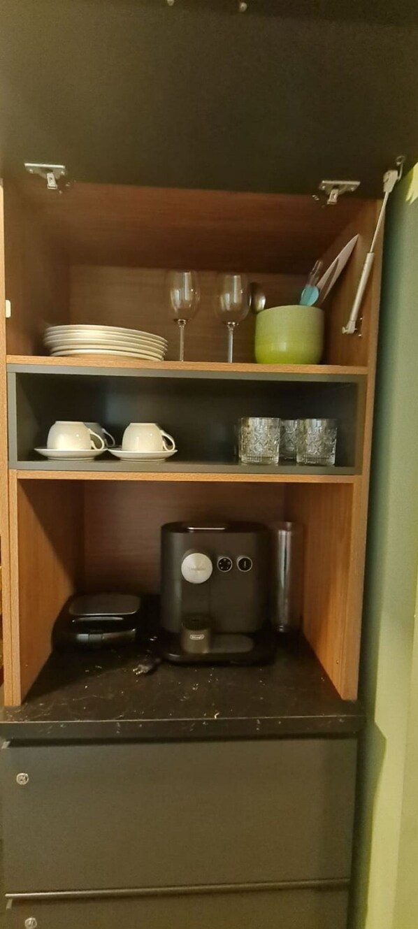 A kitchen with a coffee maker, a shelf with plates, cups, and glasses, and a green bowl. - CJC Short Stay Ilusion (Natal)