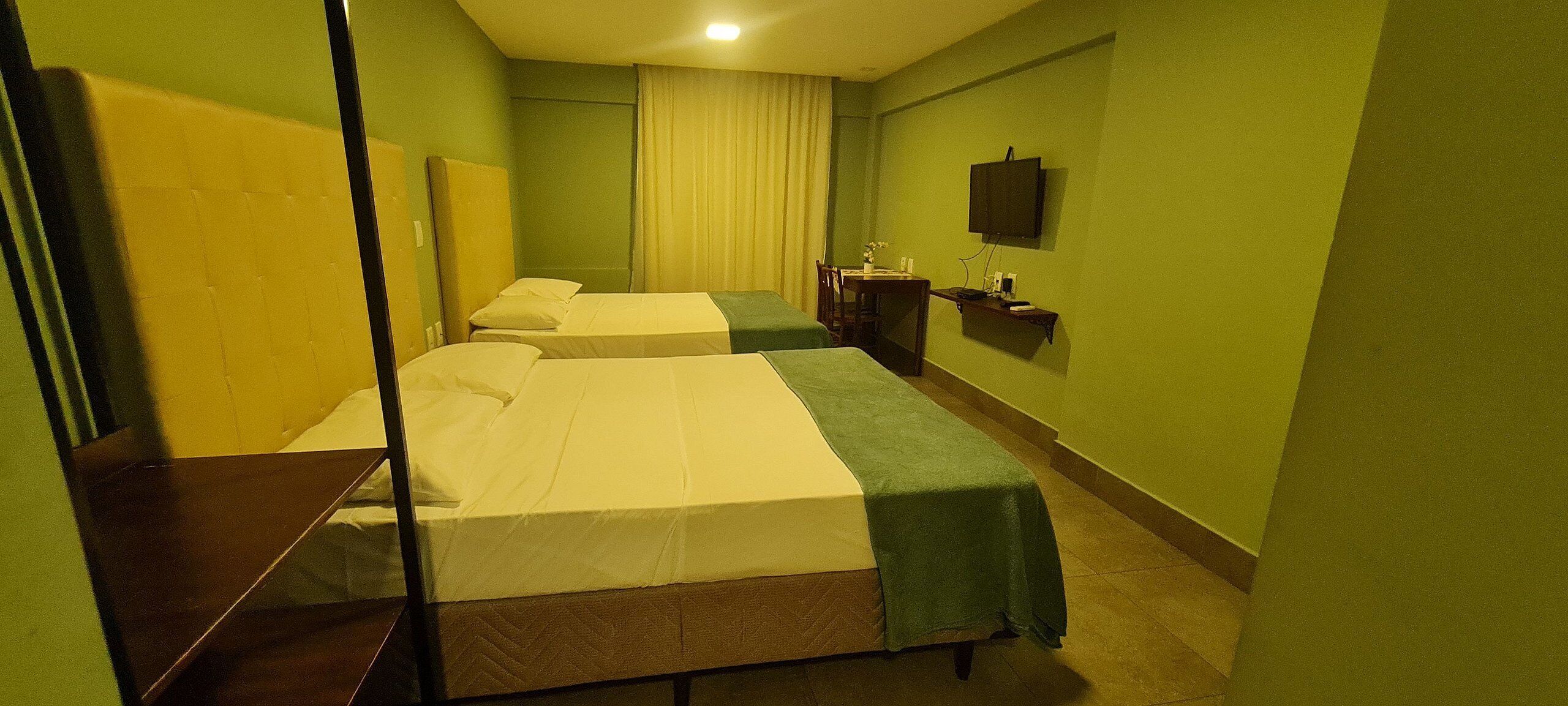 Quarto de hotel com duas camas, uma escrivaninha, uma cadeira, uma televisão e uma janela com cortinas.